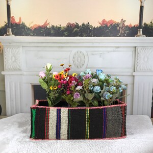 Op de afbeelding: Een zwart-wit gestreepte geweven mand met een roze, rode, gele, blauwe en groene rand. De mand is gevuld met kleurrijke kunstbloemen.
