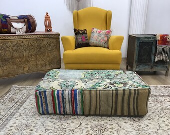 May include: A yellow armchair with two patterned cushions sits in front of a wooden chest and a blue cabinet. A large, square, multi-coloured, patterned footstool sits in front of the chair on a patterned rug.