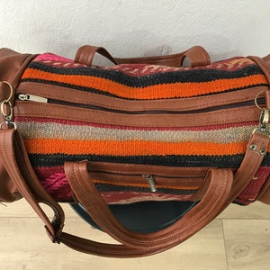 Op de afbeelding: Een bruine leren reistas met een kleurrijk gestreept patroon. De tas heeft twee bovenste handvatten en een schouderband.