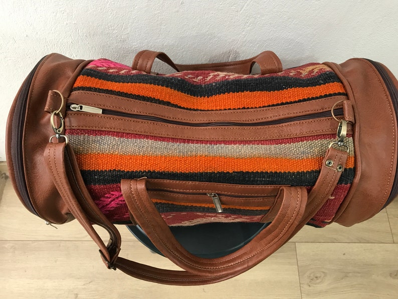 Op de afbeelding: Een bruine leren reistas met een kleurrijk gestreept patroon. De tas heeft twee bovenste handvatten en een schouderband.