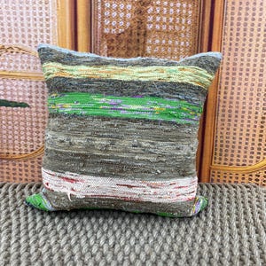 May include: Square decorative pillow with a textured, woven design. The pillow features horizontal stripes in shades of green, gray, and white. The pillow is placed on a woven surface.