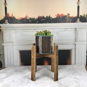 Può includere: Un portavasi in legno marrone con una pianta in vaso in un vaso a strisce marroni e bianche. Il portavasi è davanti a un camino in marmo bianco con una ghirlanda di verde sopra.