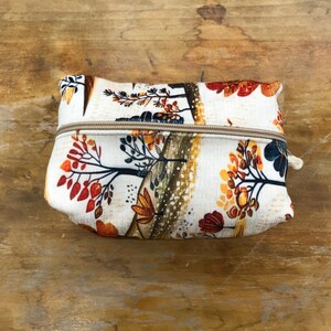 May include: A rectangular zipped pouch featuring an off-white fabric base with a printed pattern of stylised trees and foliage in shades of orange, red, brown, and dark blue. The pouch has a beige zip closure running across the top centre.