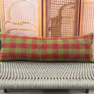 May include: A rectangular pillow with a red and green plaid pattern. The pillow is resting on a woven bench. The background includes a wooden screen and a green plant.