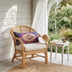 May include: A natural rattan chair with a cream cushion and a decorative pillow with a purple, blue, and orange pattern. A straw hat rests on the chair. A small white table holds a book, a glass of water, and a bowl of fruit.