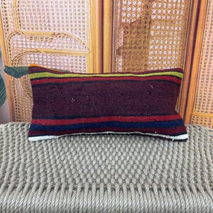 May include: A rectangular throw cushion featuring a woven textile cover with horizontal stripes in deep burgundy, red, navy blue, yellow, and green. The fabric has a textured appearance with visible thread details and a white border along the bottom edge.