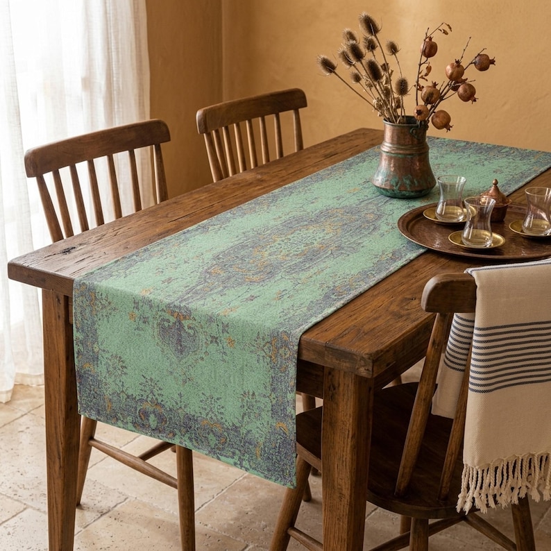 a wooden dining table with a green and blue patterned runner, surrounded by wooden chairs. On the table, there is a vase with dried flowers and a tray with cups and saucers.