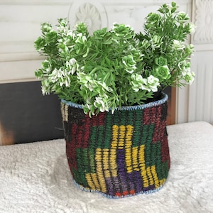 May include: A cylindrical fabric planter featuring a woven geometric pattern in shades of dark red, forest green, and mustard yellow. The top and bottom edges are finished with a light blue stitched trim. A lush green artificial plant sits inside.