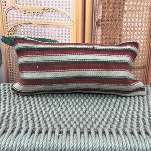 May include: A rectangular cushion with horizontal stripes in red, brown, and white. It rests on a grey woven surface. The background includes a woven wooden panel and green leaves.