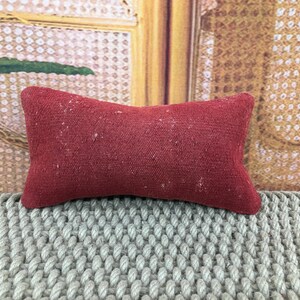 May include: A small, rectangular, red cushion with a textured surface. The cushion is placed on a grey, woven surface. The background features a decorative wooden panel with a patterned design.