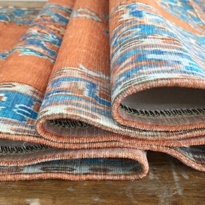 May include: Close-up of a folded orange and blue patterned rug with a fringed edge. The rug is folded in thirds, revealing the underside of the fabric.