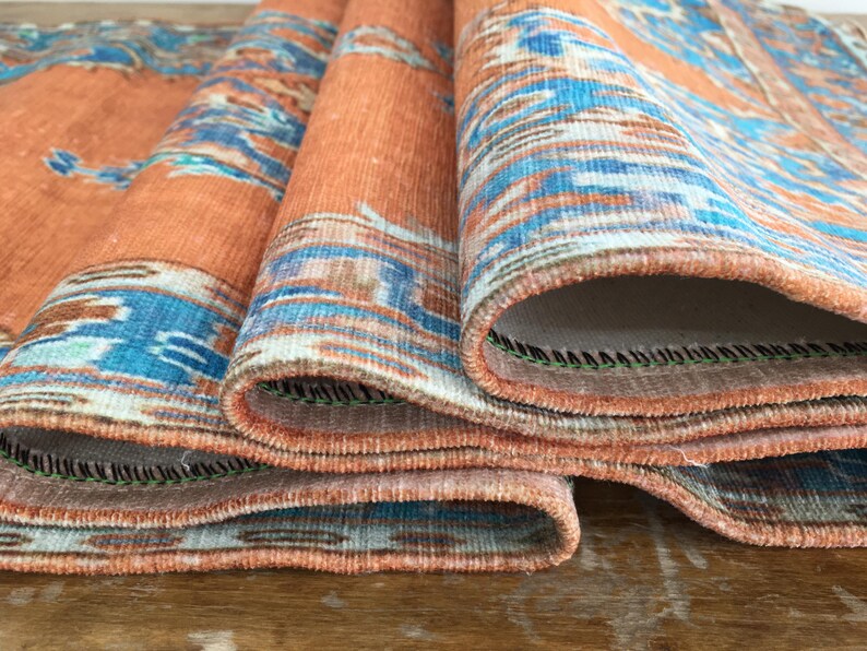 May include: Close-up of a folded orange and blue patterned rug with a fringed edge. The rug is folded in thirds, revealing the underside of the fabric.