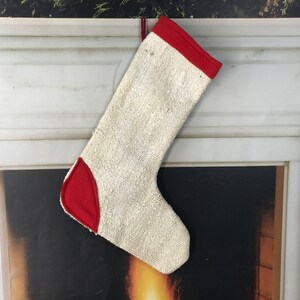 Może przedstawiać: Biała choinkowa skarpetka z czerwonymi lamówkami i czerwoną łatą na palcu. Skarpetka wisi na białym marmurowym kominku.