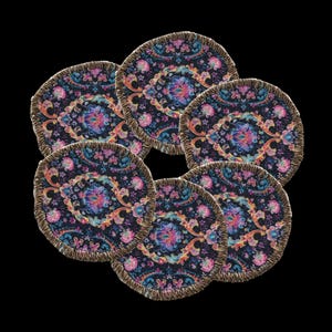 May include: Six round coasters with a floral pattern in shades of pink, blue, and purple on a dark background. Each coaster has a gold fringe edge. The coasters are arranged in a circular pattern.