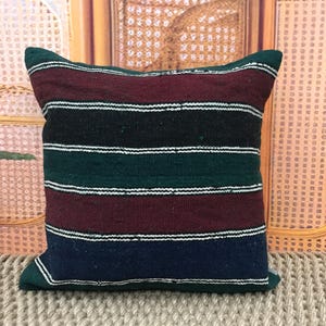 May include: Square pillow with a striped pattern in shades of burgundy, black, green, and navy blue, accented with white stripes. The pillow is made of a woven fabric and has a soft, textured appearance. The pillow is a decorative home accent.