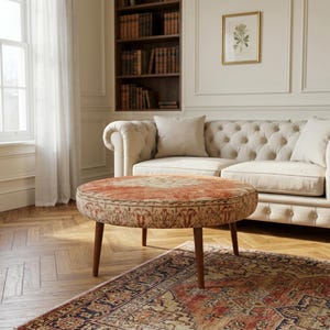 May include: A round ottoman featuring a patterned fabric top in shades of orange, cream, and brown, supported by four tapered wooden legs. The circular piece has a low profile and a flat surface, showcasing a detailed textile design with intricate borders.