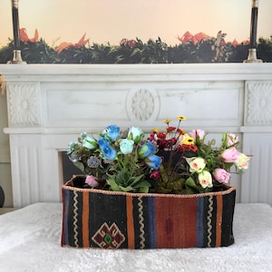 May include: A rectangular woven fabric basket featuring vertical stripes in black, orange, and blue with a geometric diamond pattern. The basket is filled with an arrangement of artificial flowers in shades of blue, pink, yellow, and deep red.