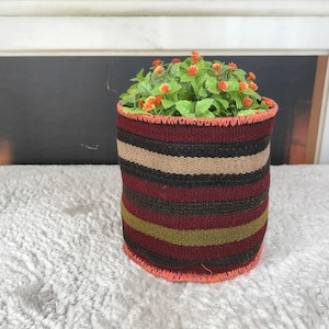 Puede incluir: Una cesta cilíndrica de tela tejida con rayas horizontales en tonos negro, rojo oscuro, verde oliva y beige. Los bordes superior e inferior están rematados con un borde cosido de color naranja brillante y contiene una pequeña planta verde con flores naranjas.
