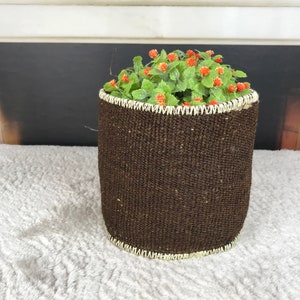 Könnte beinhalten: Ein brauner geflochtener Pflanzer mit einem weißen, gesteppten Rand, gefüllt mit einer grünen Pflanze mit kleinen orangefarbenen Blüten.