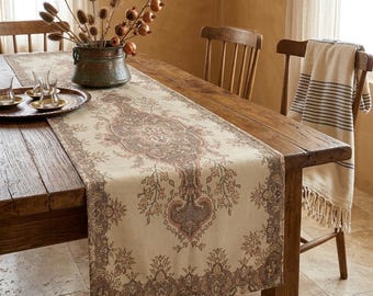 Op de afbeelding: Een beige en bruin patroon tafelloper met een bloemenontwerp wordt weergegeven op een rustieke houten tafel. De loper heeft een centraal medaillon en randdetails. Een koperen vaas met gedroogde planten en een dienblad met glazen staan ook op de tafel.