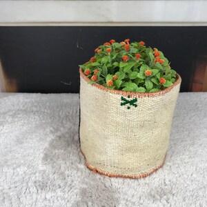 May include: A cylindrical woven planter with a cream-colored textured exterior, featuring a small green embroidered cross detail near the top rim and a thin orange stitched border along the top and bottom edges, containing a green leafy plant.
