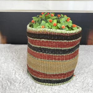 Puede incluir: Una cesta tejida cilíndrica con rayas horizontales en tonos negro, rojo óxido y beige. La cesta tiene un borde cosido de color verde claro y contiene una pequeña planta artificial con hojas verdes y diminutos capullos de flores de color naranja brillante.