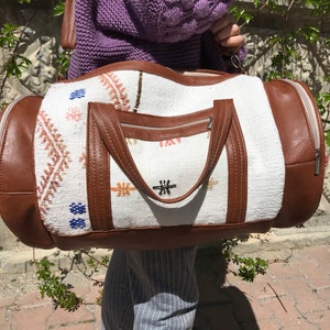 Puede incluir: Bolsa de viaje de lona blanca y marr&oacute;n con patrones geom&eacute;tricos bordados. La bolsa tiene cierre de cremallera y un asa superior.