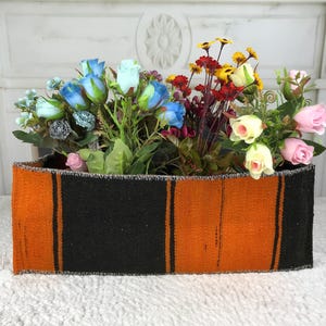 May include: A rectangular fabric basket featuring alternating vertical stripes of burnt orange and black. The basket is filled with an assortment of artificial flowers, including blue roses, pink roses, cream roses, and small yellow and red wildflowers.