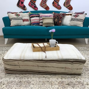 May include: A white and brown striped ottoman with a wooden tray on top. The tray has a book, a string of beads, and a white mug on it. The ottoman is sitting on a patterned rug in front of a teal blue sofa with cushions.