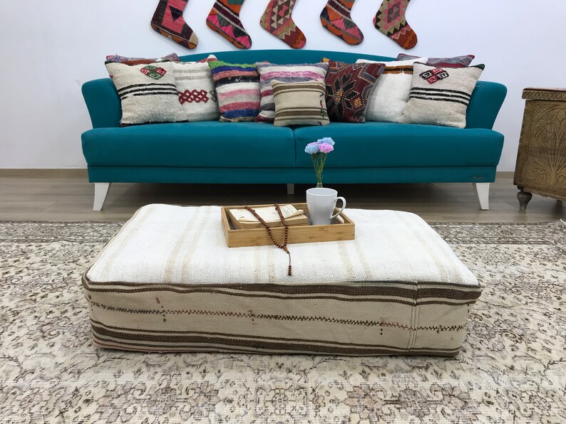 May include: A white and brown striped ottoman with a wooden tray on top. The tray has a book, a string of beads, and a white mug on it. The ottoman is sitting on a patterned rug in front of a teal blue sofa with cushions.