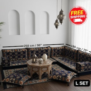 Peut inclure: Un ensemble de sièges en forme de L avec un motif bleu marine, orange et or. L'ensemble comprend des coussins, un tapis et une table basse. La section la plus longue mesure 250 cm, et la plus courte mesure 180 cm. Des lanternes décoratives sont suspendues au plafond.