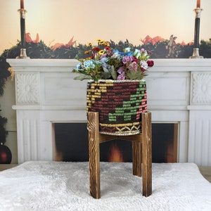 May include: A colorful woven planter with a geometric pattern sits on a wooden stand in front of a white fireplace. The planter is filled with a bouquet of pink, purple, and blue flowers.