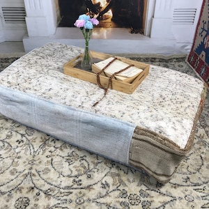 May include: A light blue and beige floor cushion with a floral pattern. The cushion is sitting on a patterned rug with a wooden tray and a book on top. A string of wooden beads is on the tray.