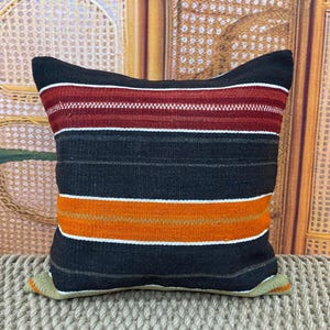 May include: A square cushion with a striped design. The cushion has horizontal stripes in black, red, white, and orange. The cushion is placed on a woven surface, and the background has a woven pattern.