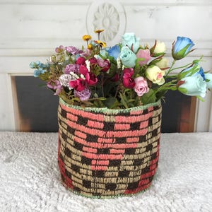 May include: A cylindrical woven basket featuring a geometric pattern in shades of dark brown, tan, and coral pink, with a light green stitched rim. The basket is filled with an assortment of artificial flowers in blue, pink, yellow, and cream.