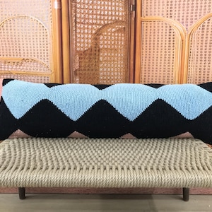 May include: A long, rectangular knitted pillow with a black and light blue zigzag pattern. The pillow is resting on a woven bench with wooden legs. The background features a wooden cabinet.