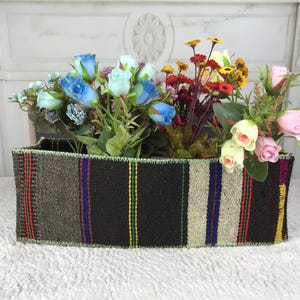 May include: A rectangular woven fabric basket featuring vertical stripes in black, brown, beige, and thin accents of red, blue, green, and yellow, filled with an assortment of artificial flowers including blue, pink, and yellow roses.