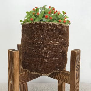 May include: A dark brown woven fabric planter with a textured surface and a light-colored stitched rim, holding a green plant with small orange flowers, supported by a four-legged wooden stand with a visible grain and dark brown finish.