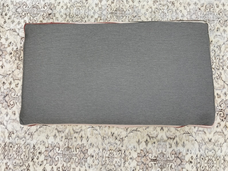 May include: A grey rectangular cushion with a dark grey fabric cover. The cushion is sitting on a patterned rug.