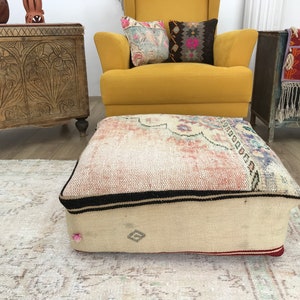 May include: A large, square, cream-colored floor cushion with a colorful, patterned top. The cushion has a black trim and a red border.