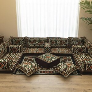 May include: A modular floor seating set featuring a U-shaped arrangement of cushions with a dark brown base and a geometric pattern in orange, green, and white. The set includes backrests, armrests, a central square ottoman, and two floor pillows.