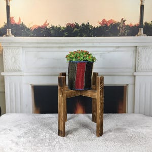 May include: A dark brown wooden plant stand with a cross-shaped base and four vertical legs, supporting a cylindrical planter. The planter features a woven fabric exterior with vertical stripes in black, red, and blue, containing a small green plant.