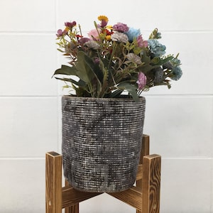 Könnte beinhalten: Ein zylindrischer Übertopf mit dunkelgrauem und weißem Webmuster, gefüllt mit einem Strauß künstlicher Blumen in Rosa, Blau, Gelb und Violett, auf einem vierbeinigen Holzständer mit sichtbarer Maserung und brauner Oberfläche stehend.