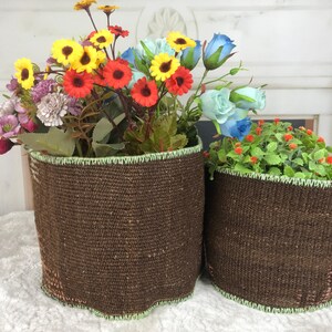 May include: Two cylindrical brown woven baskets with a light green stitched trim along the top and bottom edges. The larger basket is filled with a colorful arrangement of yellow, red, and blue artificial flowers, while the smaller holds greenery.