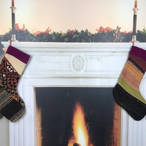 May include: Two colorful Christmas stockings hang on a white marble fireplace mantel. The stockings are made of woven fabric with a variety of patterns and colors, including red, green, brown, and black. The fireplace has a fire burning inside.
