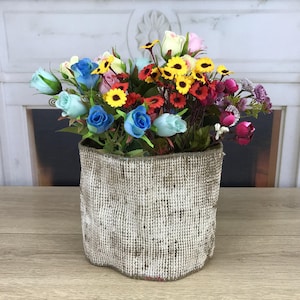 May include: A cylindrical container with a textured, beige and brown woven exterior filled with an arrangement of artificial flowers in shades of blue, yellow, red, pink, and white, featuring small sunflowers and various rose-like blossoms.