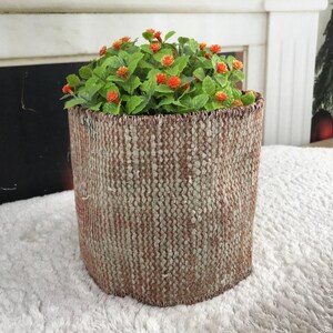 May include: A cylindrical plant pot featuring a textured, woven exterior in muted shades of beige and faded red. The pot contains a lush green plant with small, vibrant orange flowers that bloom across the top, creating a contrast in color and texture.