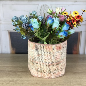 May include: A woven basket with a pink, white, and brown plaid pattern holds a bouquet of artificial flowers. The flowers are blue, pink, white, and yellow.