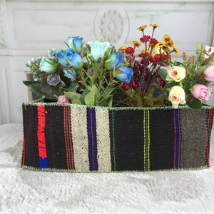 May include: A woven rectangular planter with a black background and colorful stripes in red, blue, green, and yellow. The planter is filled with artificial flowers, including blue roses, yellow daisies, and pink roses.