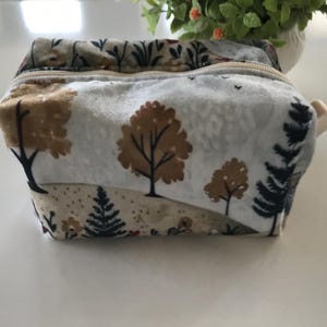 May include: A rectangular fabric pouch featuring a printed landscape pattern of brown and black trees against a light gray background. The pouch has a top zipper closure and a small light-colored tab on the side, showcasing a soft, textured surface.